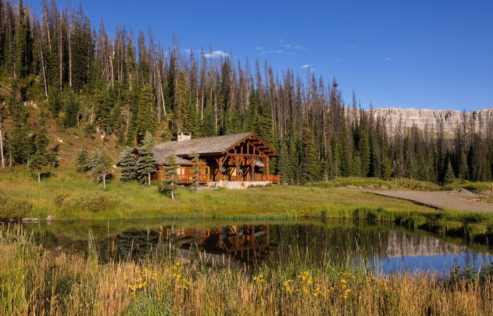 A large cabin overlooking a pond with pine trees in the background.