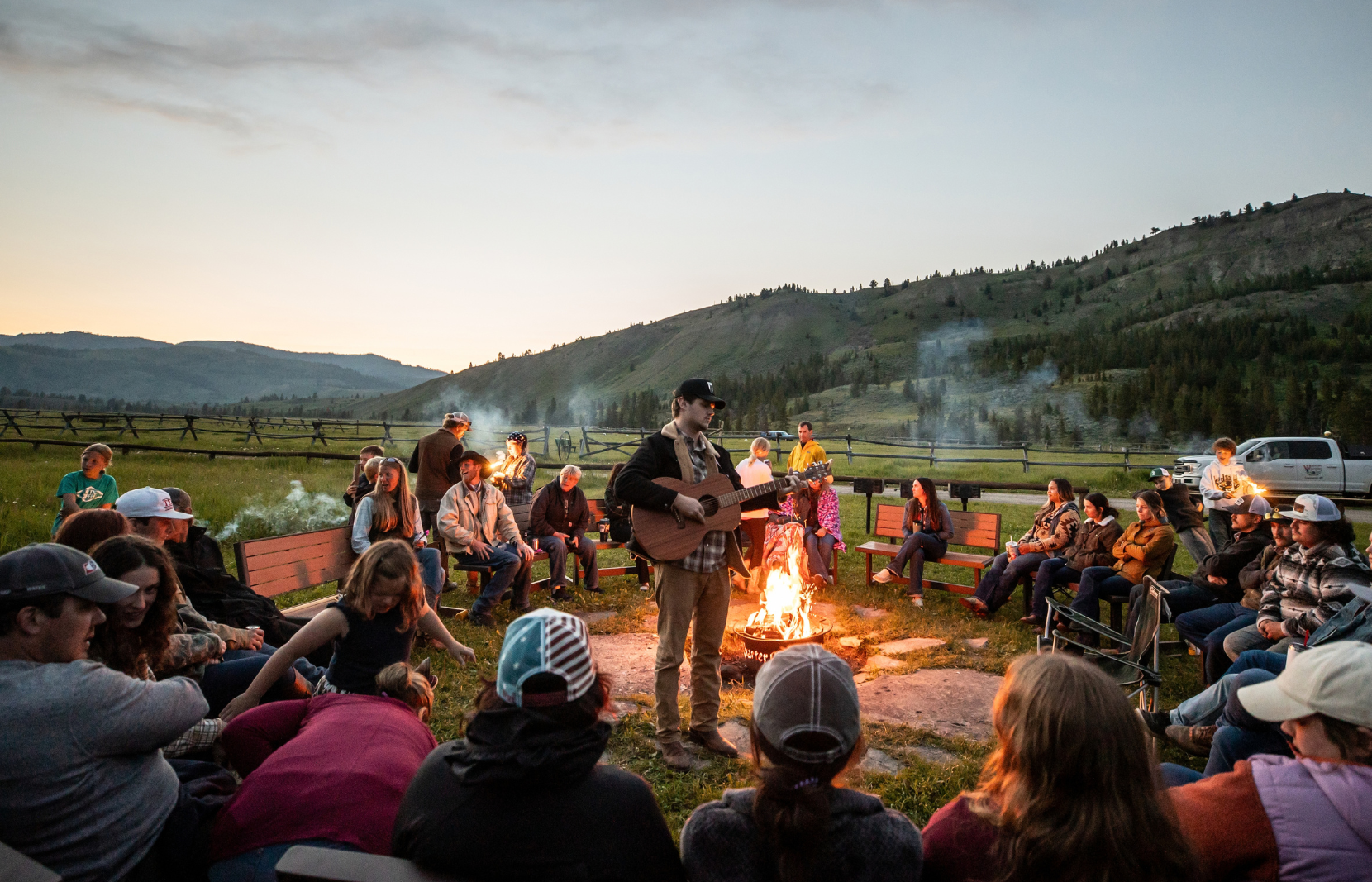 A man playing a guitar in front of a campfire to a large group of people.