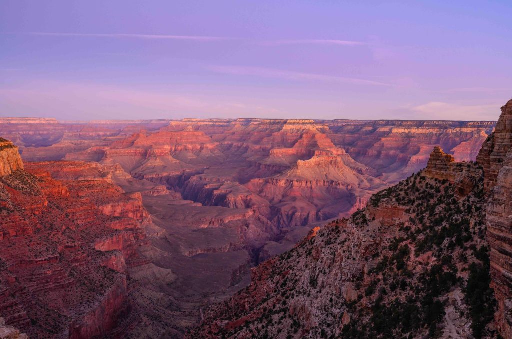 Layers of red and pink sandstone buttes and canyon walls glow in warm twilight light across the vast expanse of Grand Canyon National Park, Arizona, under a soft purple sky.