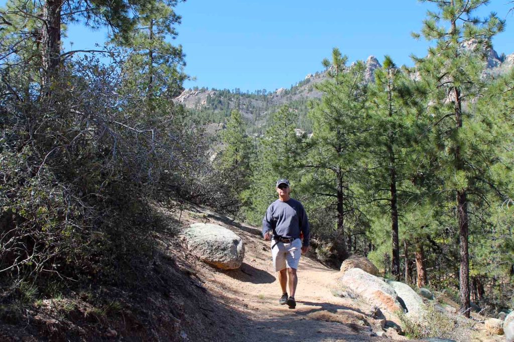 A hiker walks along a rocky dirt trail through a pine forest at Hualapai Mountain Park near Kingman, Arizona, with granite rock outcroppings and tree-covered peaks visible in the background.