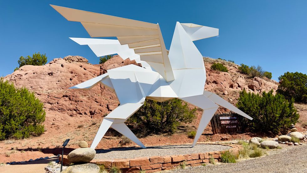 Large white, angular geometric sculpture of a winged Pegasus in mid-stride, set against red rock formations and junipers. Santa Fe County. 