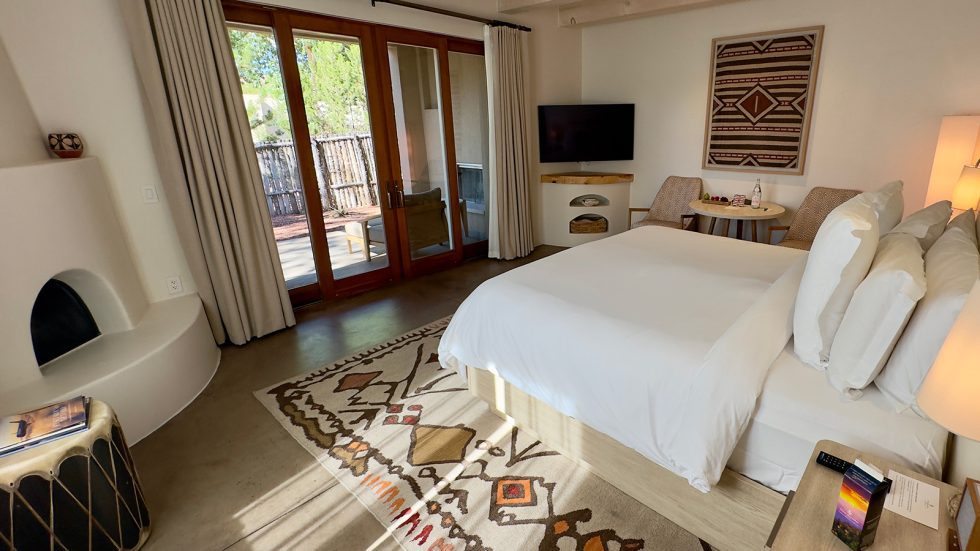 Bedroom in a Southwestern-style casita with a kiva fireplace, patterned Native-inspired rug and wall hanging, white bed, and glass doors opening onto a private patio. Santa Fe County.