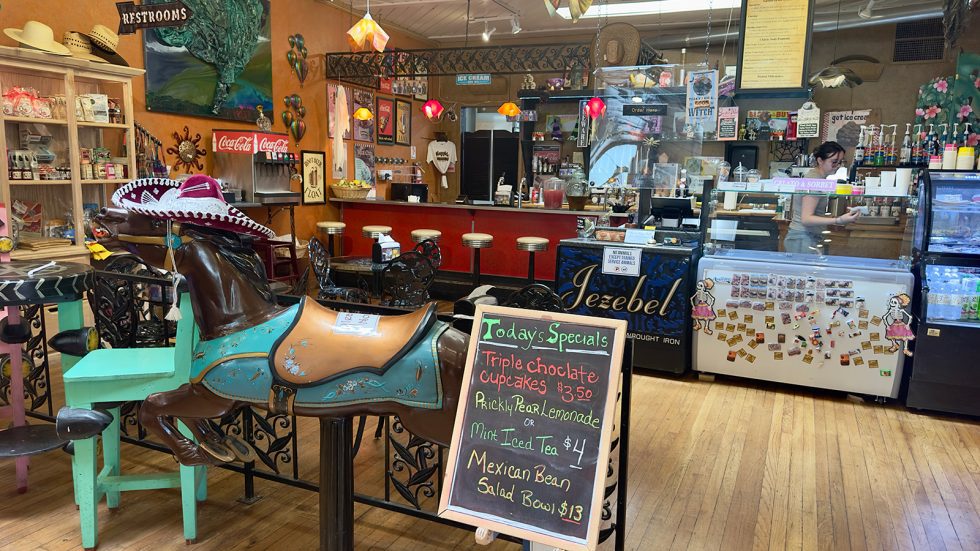 Interior of Jezebel's soda fountain and gallery with a sombrero-topped carousel horse, a chalkboard specials sign, vintage bar stools, ice cream cases, and gift shop shelves. Santa Fe County.