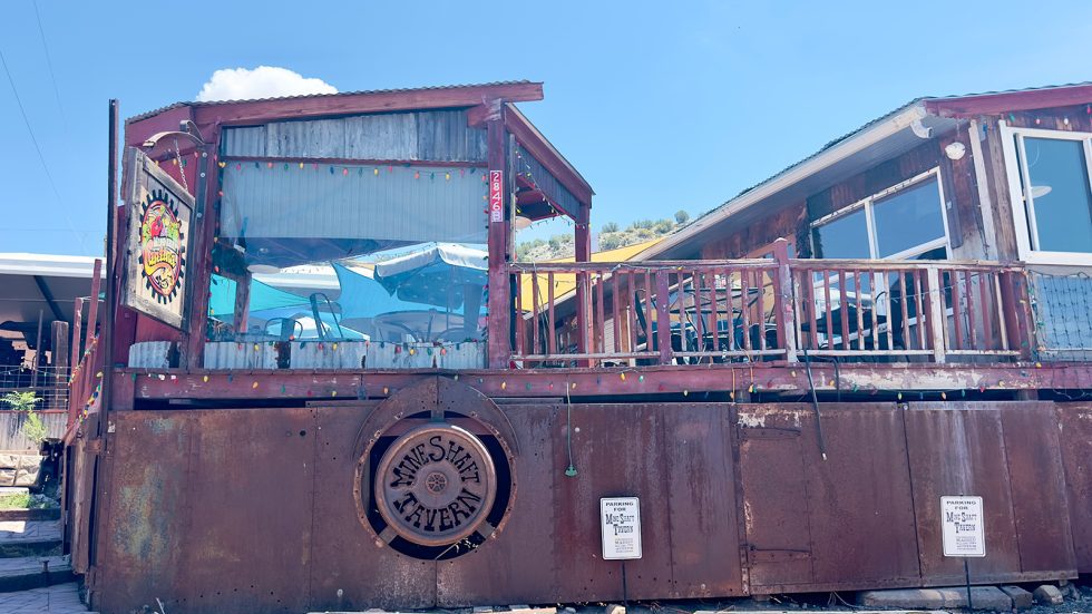 Rustic wooden building with a rusted metal facade featuring a round "Mine Shaft Tavern" sign, string lights, a covered deck with patio furniture, under a blue sky with hills behind. Santa Fe County.