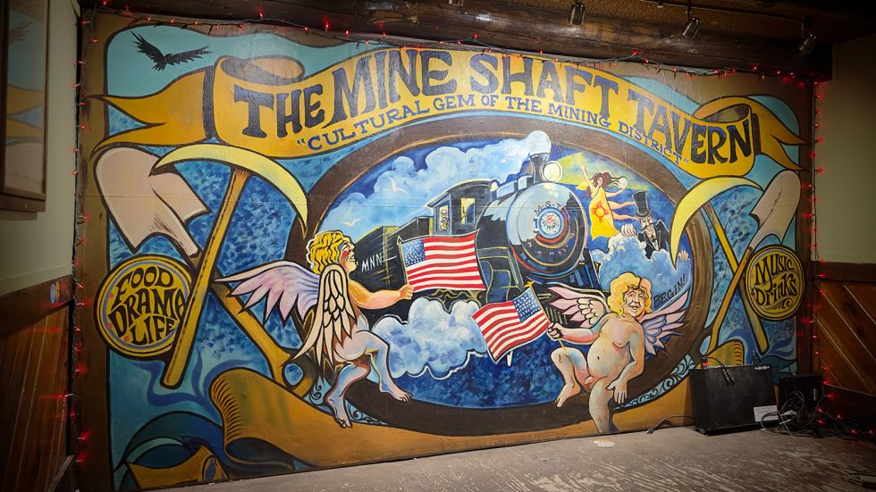 Colorful backstage mural at The Mine Shaft Tavern featuring a steam locomotive, cherubs holding American flags, crossed pickaxes, and banners reading "Food Drama Life" and "Music & Drinks." Santa Fe County.