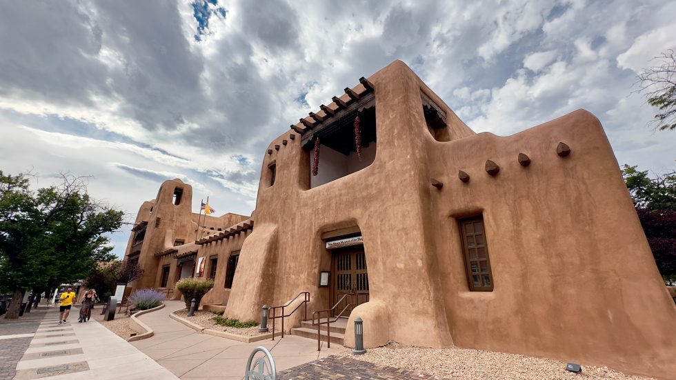 Pueblo Revival-style adobe exterior of the New Mexico Museum of Art, with rounded walls, vigas, hanging chile ristras, and wooden entrance doors, under a cloudy sky. Santa Fe County.