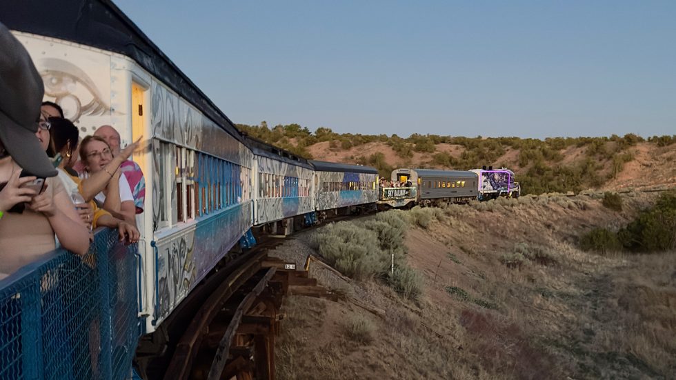 Passengers lean over an open-air rail car painted with mythical creature murals in blue, lavender, and silver, as the colorful train curves over a wooden trestle through high-desert scrubland at sunset. Santa Fe County