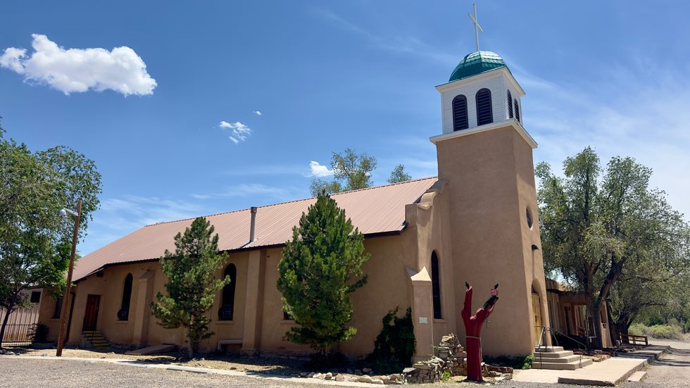100-year old adobe church with a rust-colored metal roof and a tall bell tower topped with a green dome and white cross, set among trees under a blue sky. Santa Fe County. 