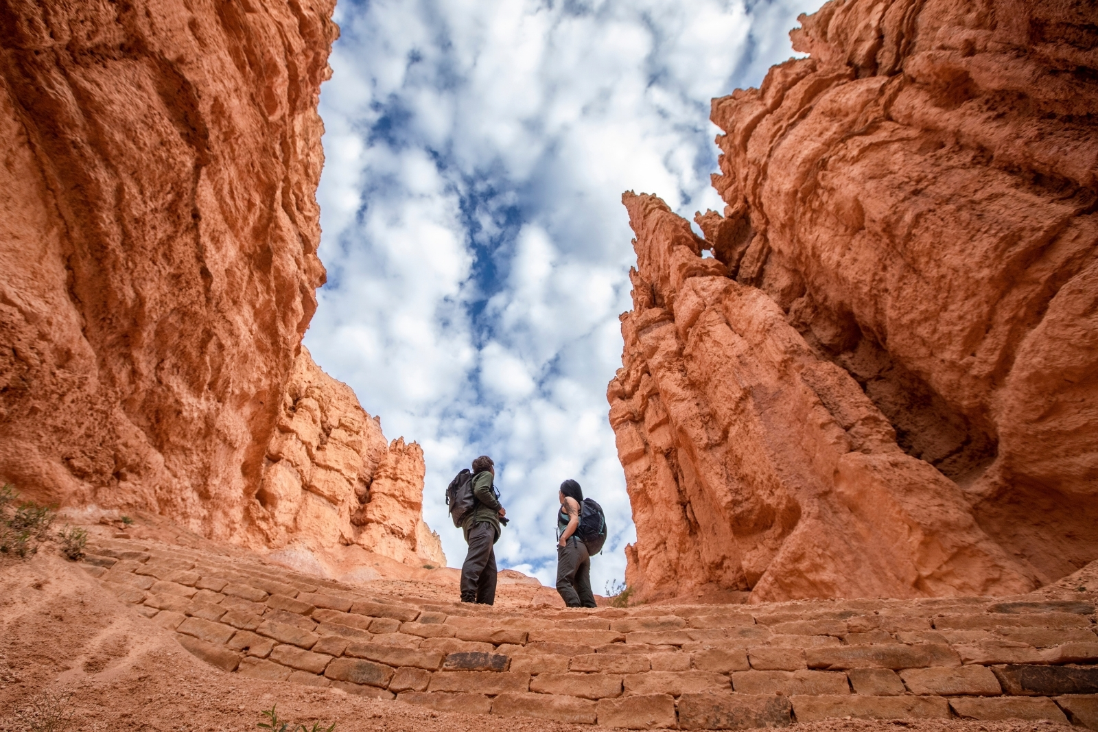 2 people in the middle of a switchback in Bryce Canyon National Park, with towering red cliffs on either side of them.